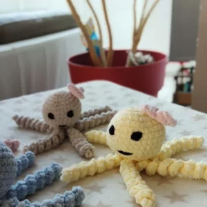 Petites pieuvres au crochet faites à la main sur une table, en beige et jaune, avec des visages noir et des oreilles roses, fond de plante d’intérieur.