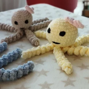 Petites pieuvres au crochet faites à la main sur une table, en beige et jaune, avec des visages noir et des oreilles roses, fond de plante d’intérieur.