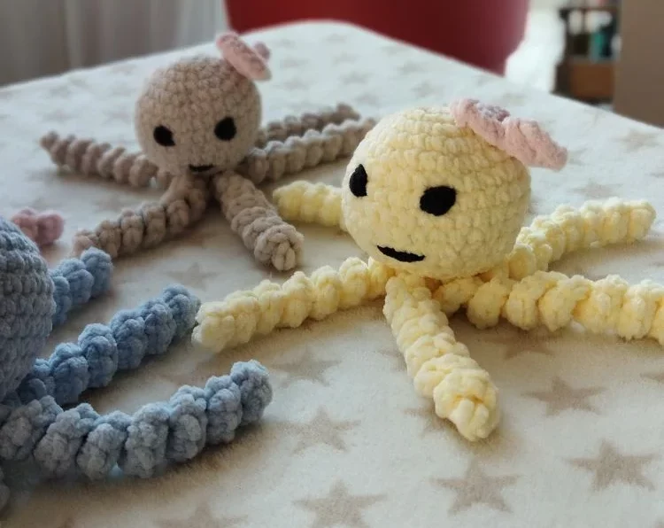 Petites pieuvres au crochet faites à la main sur une table, en beige et jaune, avec des visages noir et des oreilles roses, fond de plante d’intérieur.