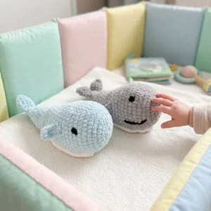 Doudou baleine au crochet, deux baleinettes pastel sur un berceau décoré, une main de bébé les approche.